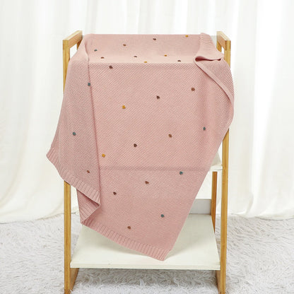 Pink knitted blanket with small colorful dots draped over a wooden chair against a white curtain background.