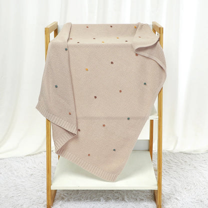 Beige knit blanket with colorful dots draped over a wooden crib against a white curtain background.