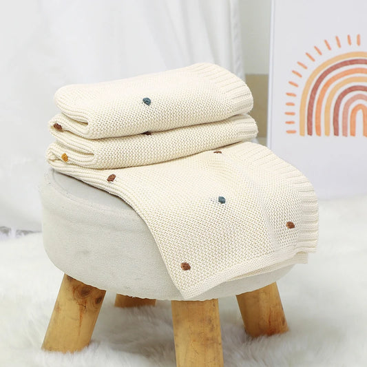 Folded blanket with small brown and blue accents on a wooden stool against a white wall with a rainbow decoration.