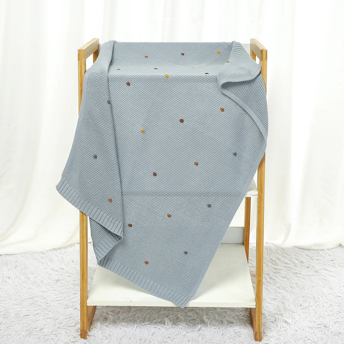 Light blue knitted blanket with small colorful dots draped over a wooden chair against a white curtain background.