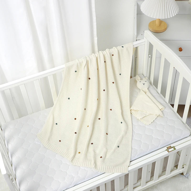 White crib with a cream-colored blanket featuring black polka dots in a nursery setting.