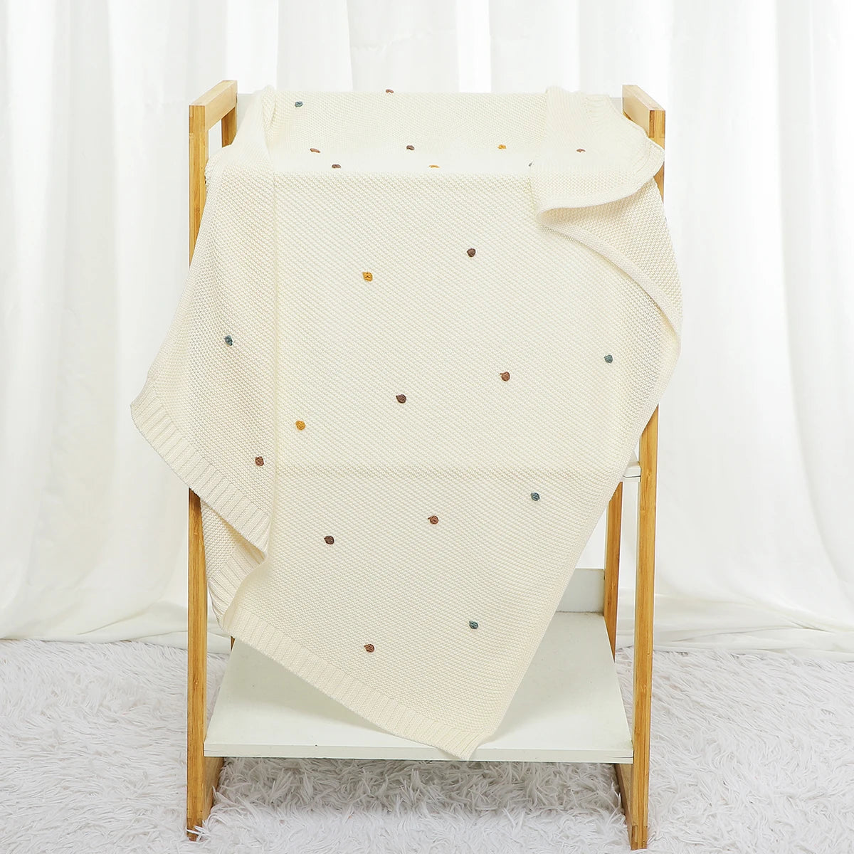 Beige blanket with colorful polka dots draped over a wooden chair against a white curtain background.