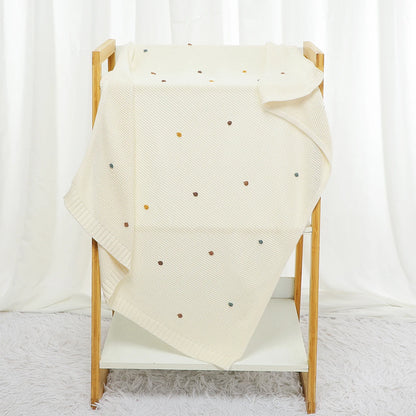 Beige blanket with colorful polka dots draped over a wooden chair against a white curtain background.